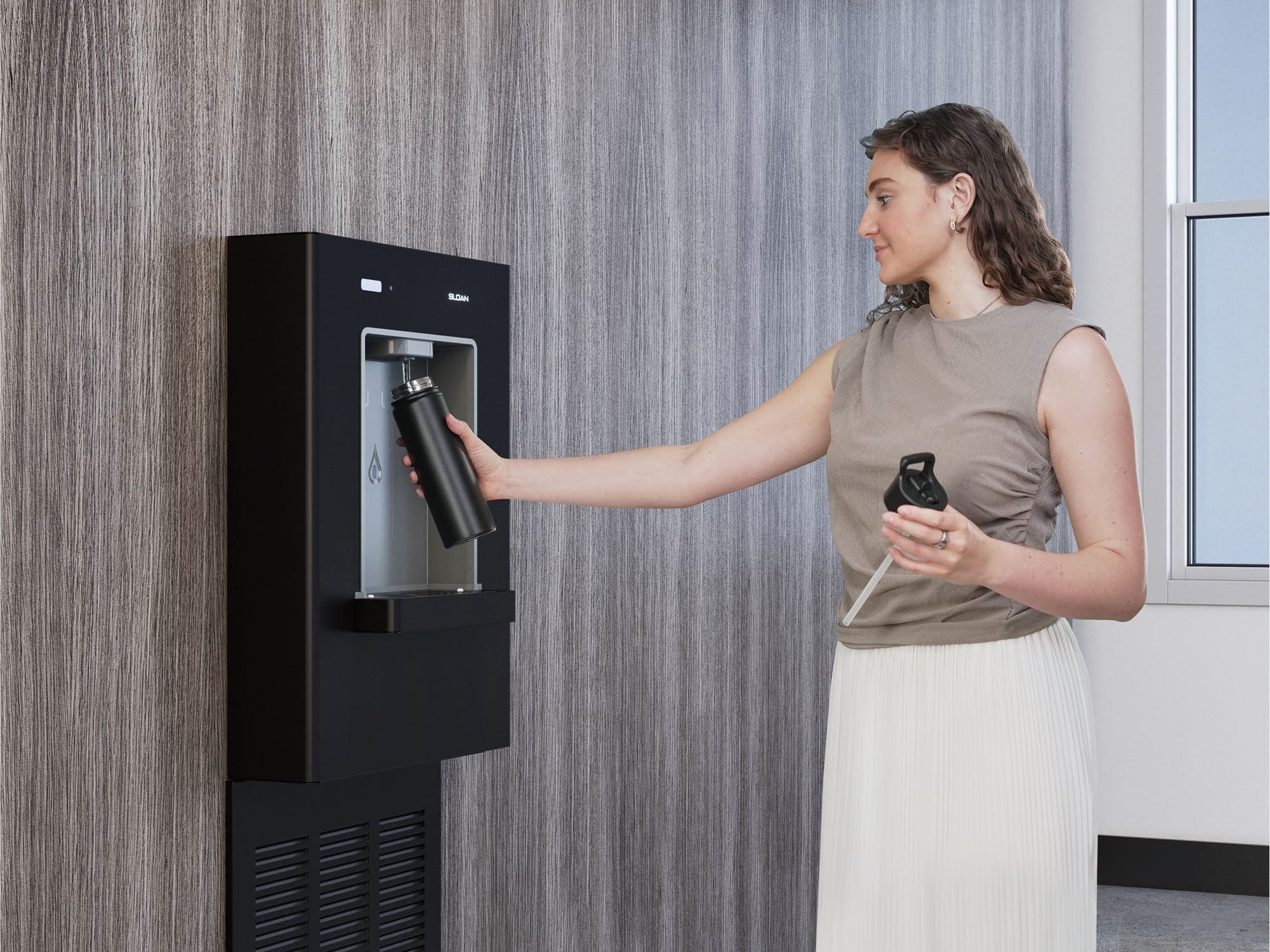 A woman fills a reusable bottle at a modern black wall-mounted water station in an office setting.