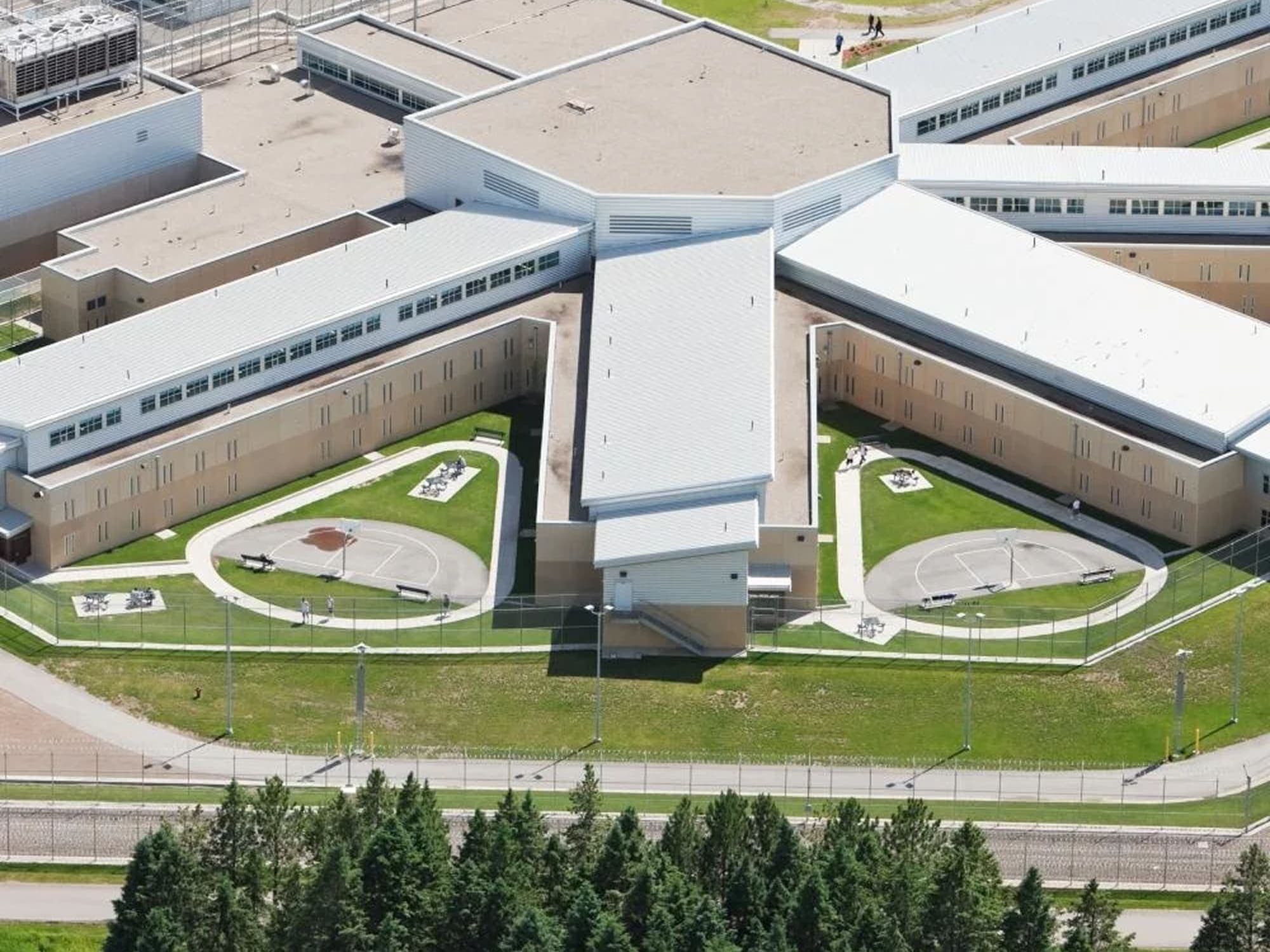 Aerial view of a large correctional facility with multiple connected wings arranged in a radial layout, surrounded by fencing and open green areas.