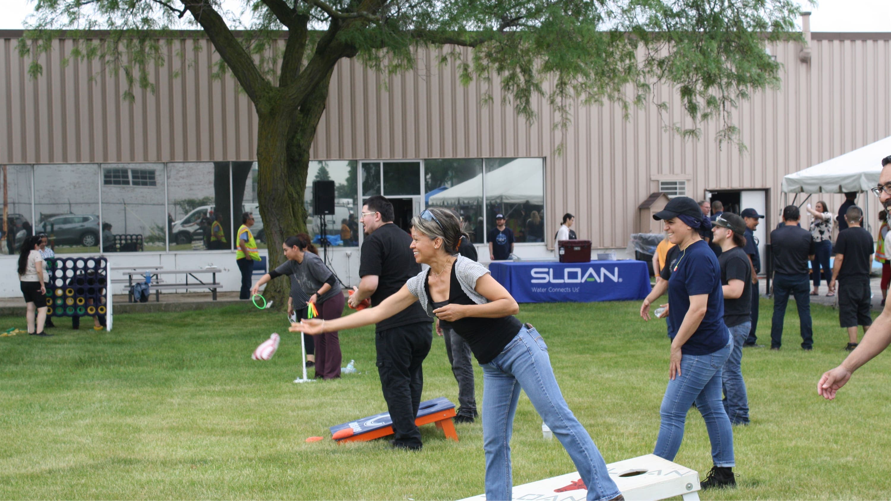 Sloan employees playing an outdoor bean bag toss game on a grassy lawn during a company event, with a Sloan banner visible in the background.