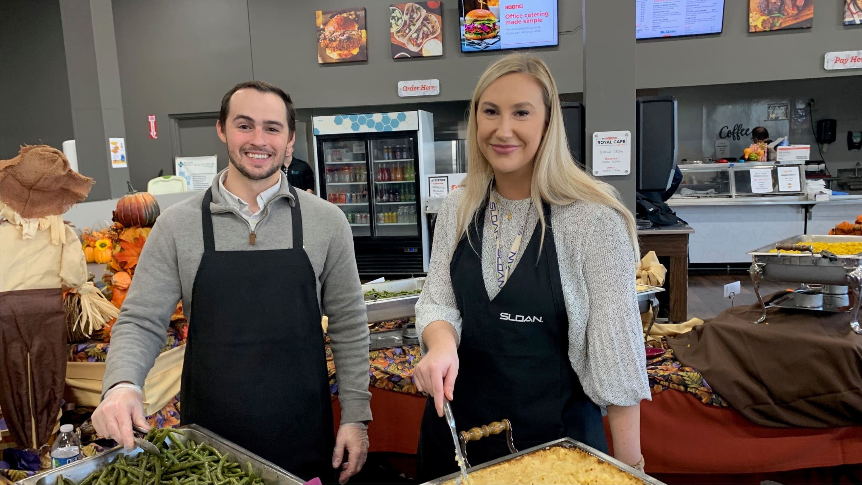 Sloan employees wearing aprons serve food from buffet trays in a cafeteria decorated with fall-themed decor.