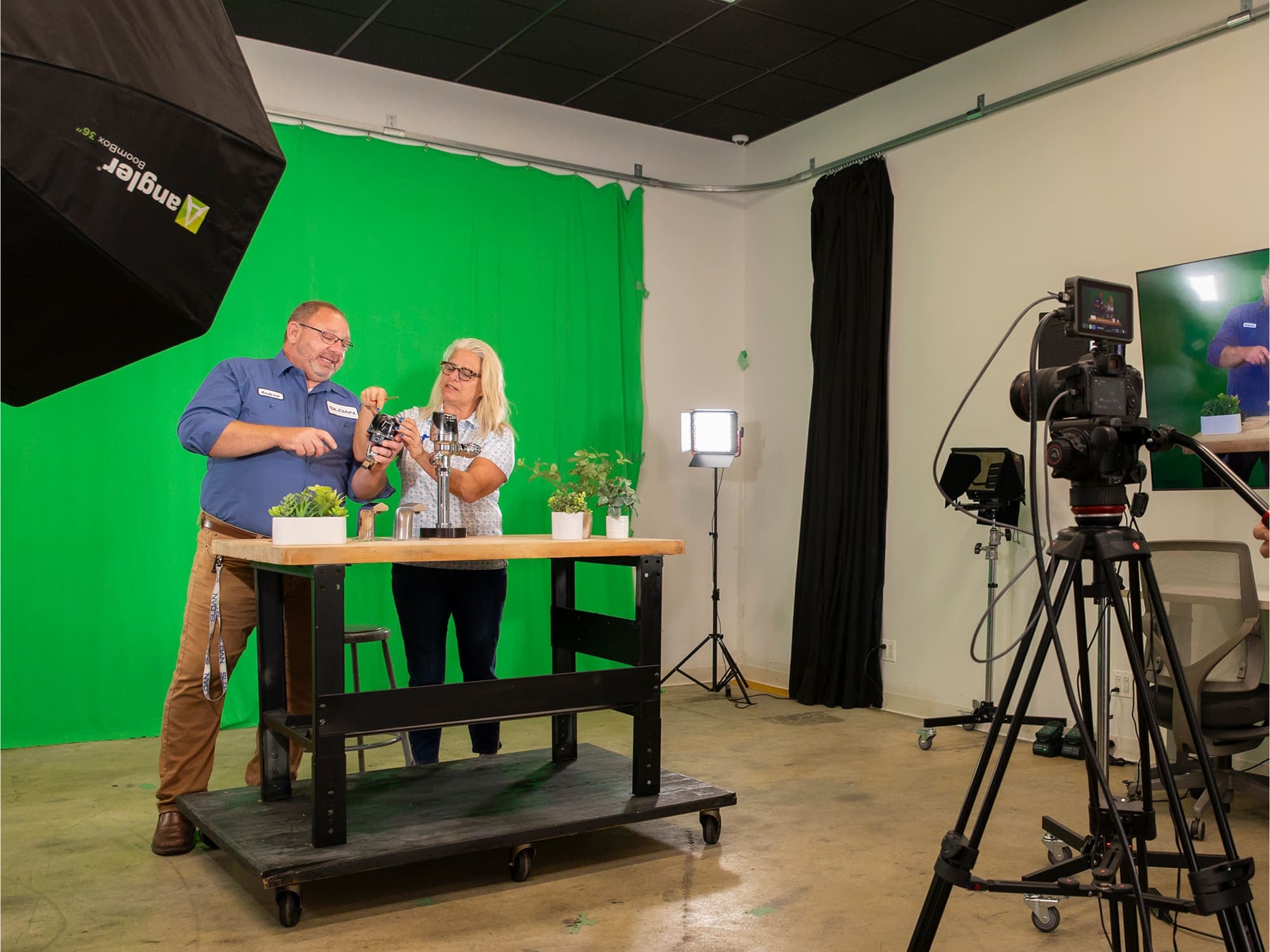 Two people standing at a table in a video production studio with a green screen background, studio lighting, and cameras set up for filming.