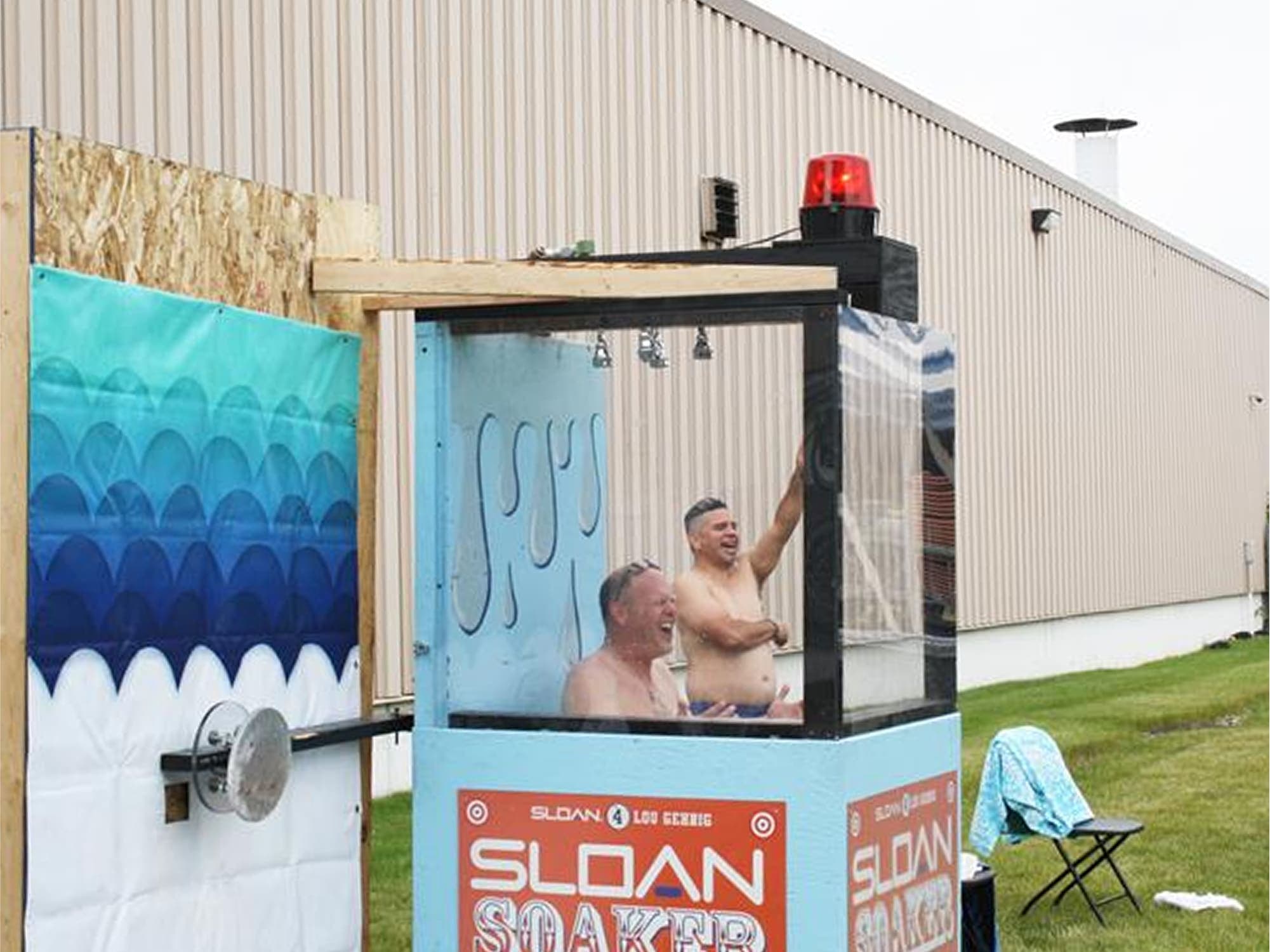 Two people sitting inside a dunk tank labeled 'Sloan' at an outdoor company event, with a warehouse building in the background.