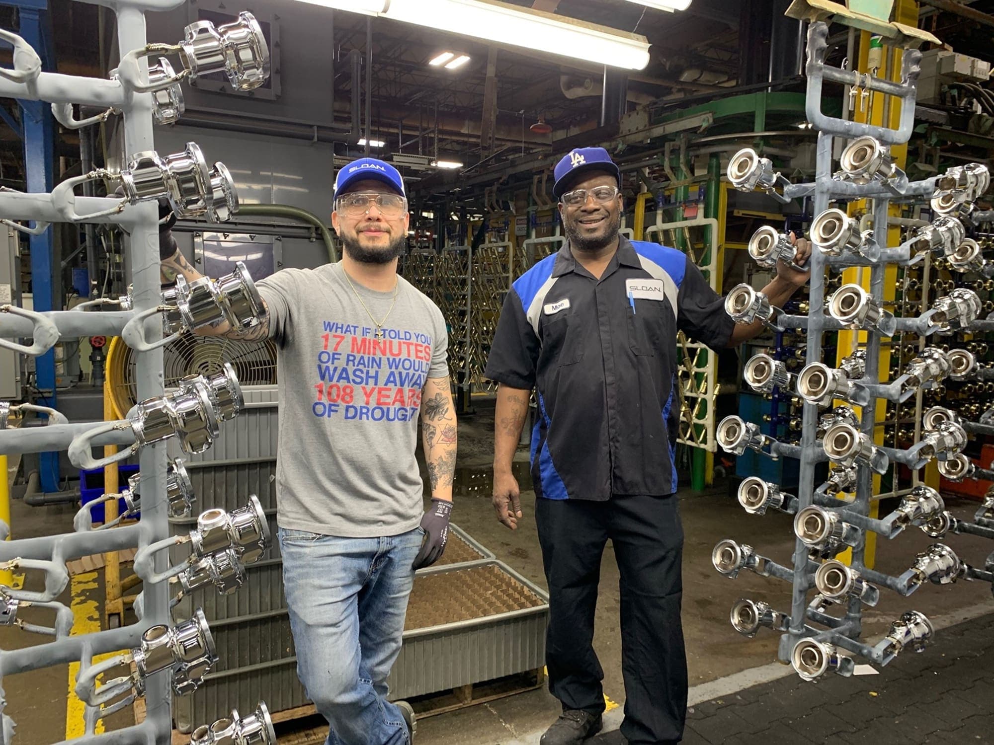 Two factory workers standing between metal racks holding polished plumbing parts inside an industrial manufacturing facility.