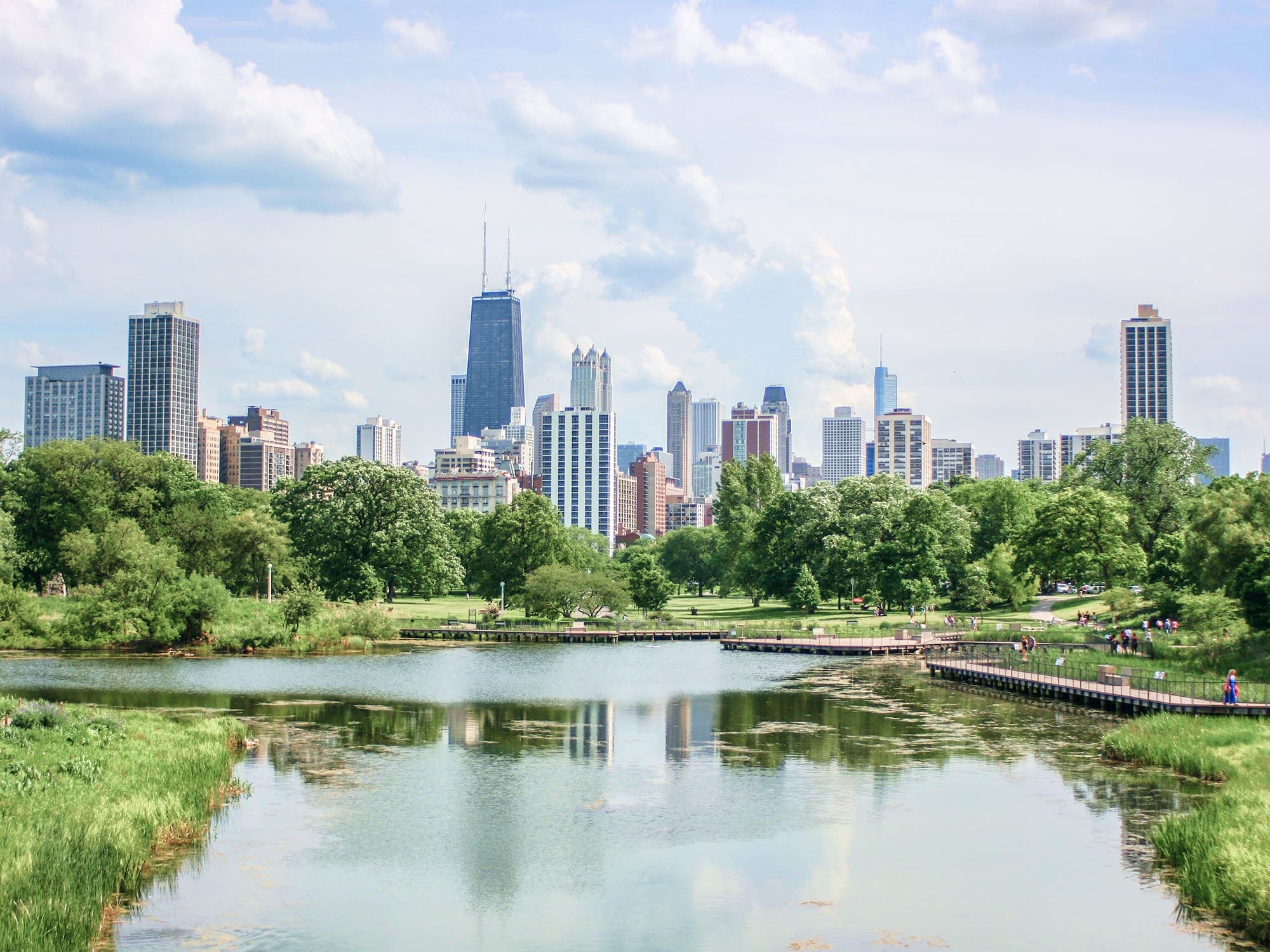 View of Chicago skyline from Lincoln Park, showing modern skyscrapers in the distance behind lush green trees and a calm reflective pond under a partly cloudy sky.