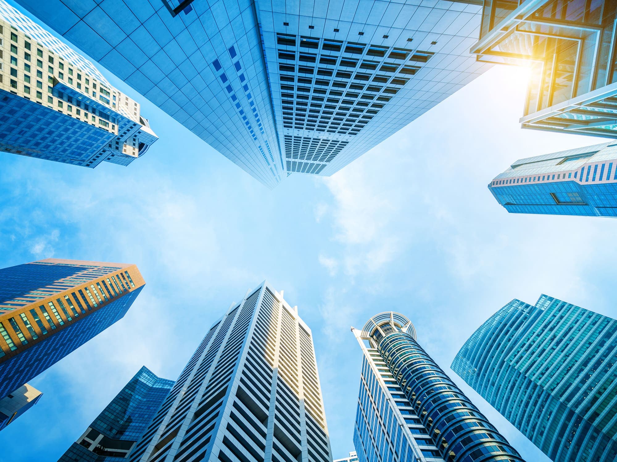 Upward view of modern skyscrapers against a bright blue sky with sunlight shining between the buildings.