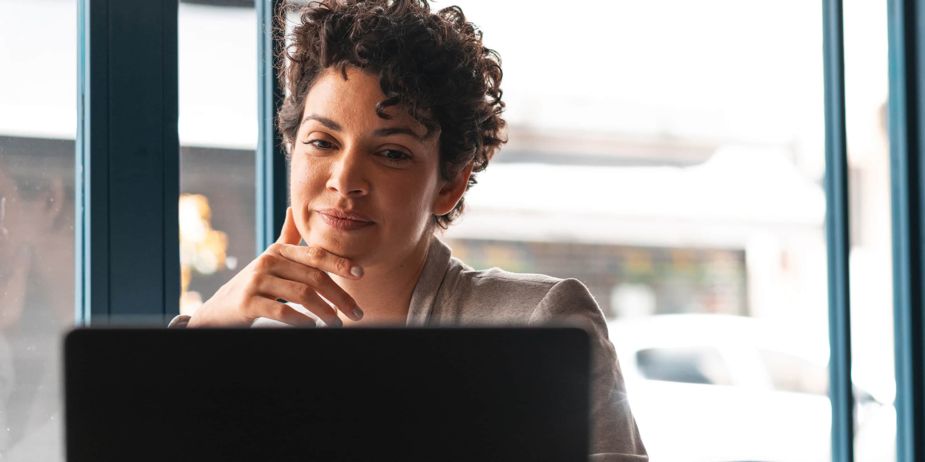 A professional woman with curly hair looking directly at the camera while working on a laptop.