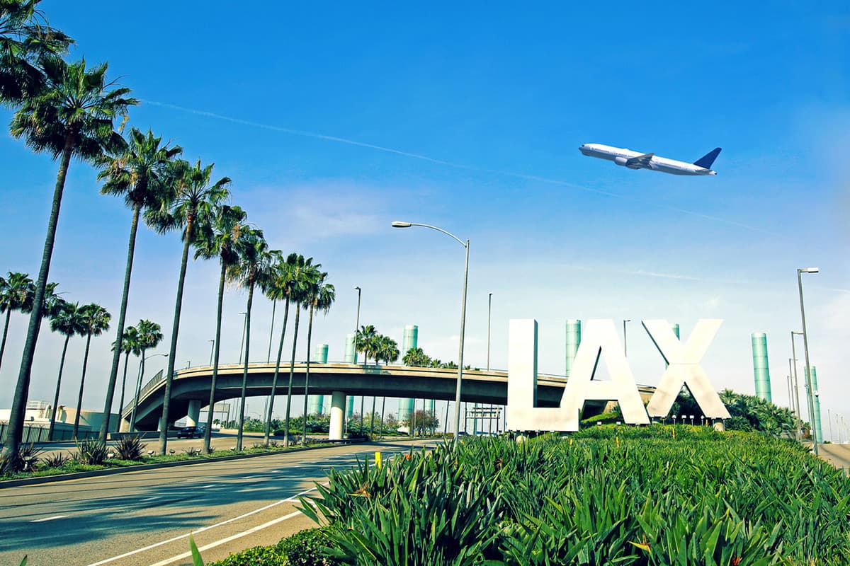 Street view of the LAX airport entrance with big letters, palm trees, greenery and an airplane flying overhead