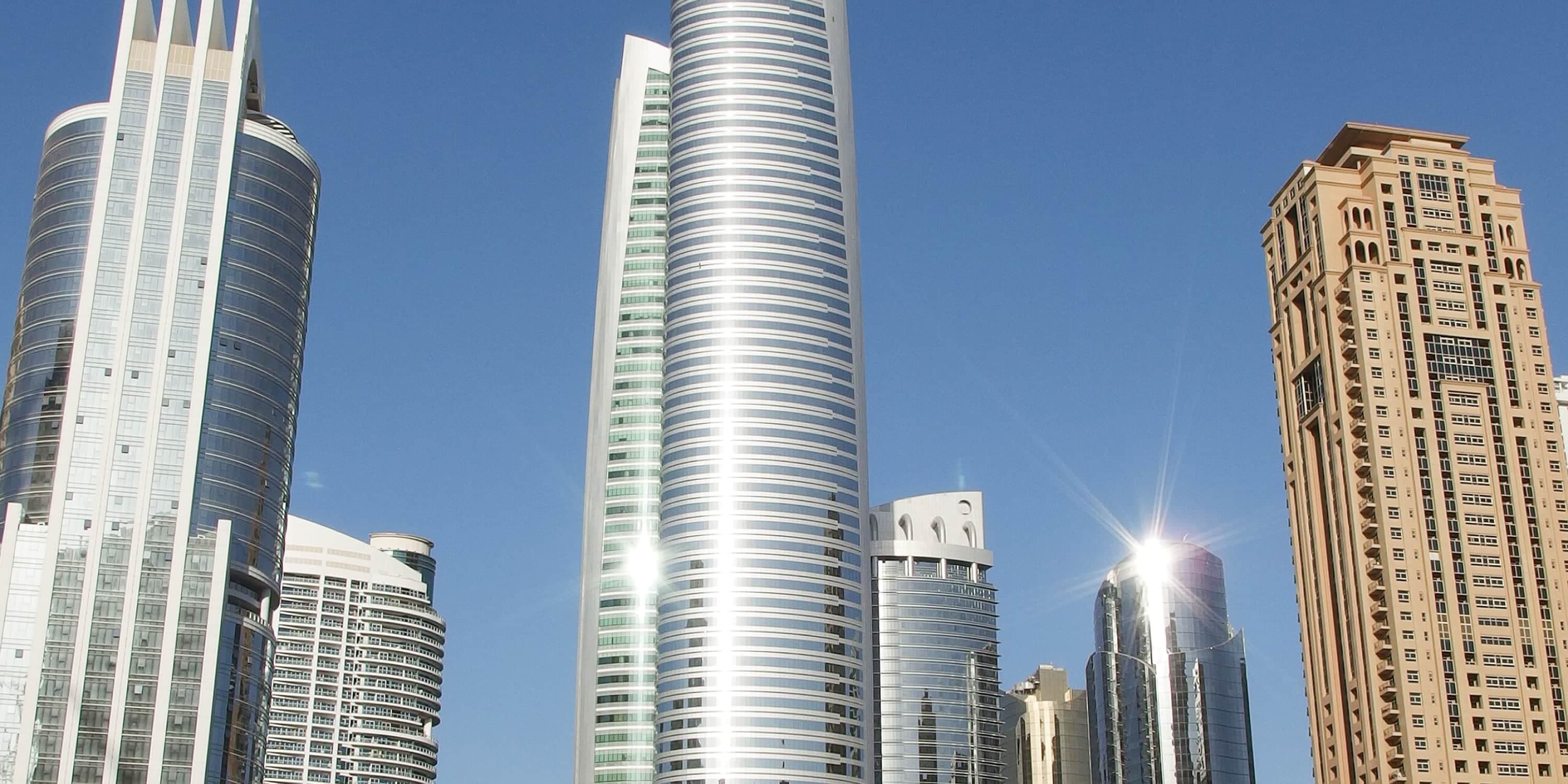 Modern skyscrapers and high-rise buildings in the Middle East, featuring glass and steel facades reflecting sunlight against a clear blue sky.