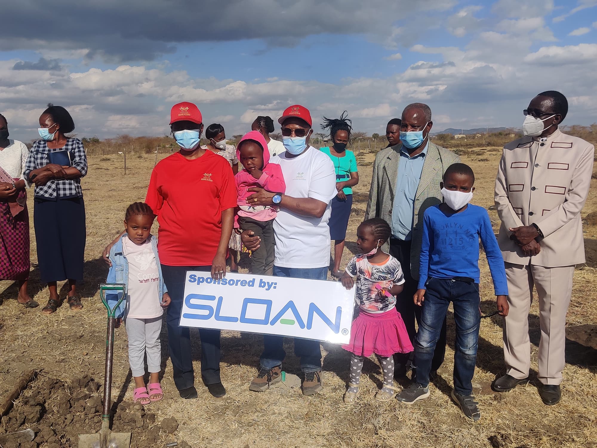 Community members and children standing together outdoors holding a sign reading Sponsored by Sloan at a Shiloh Home event