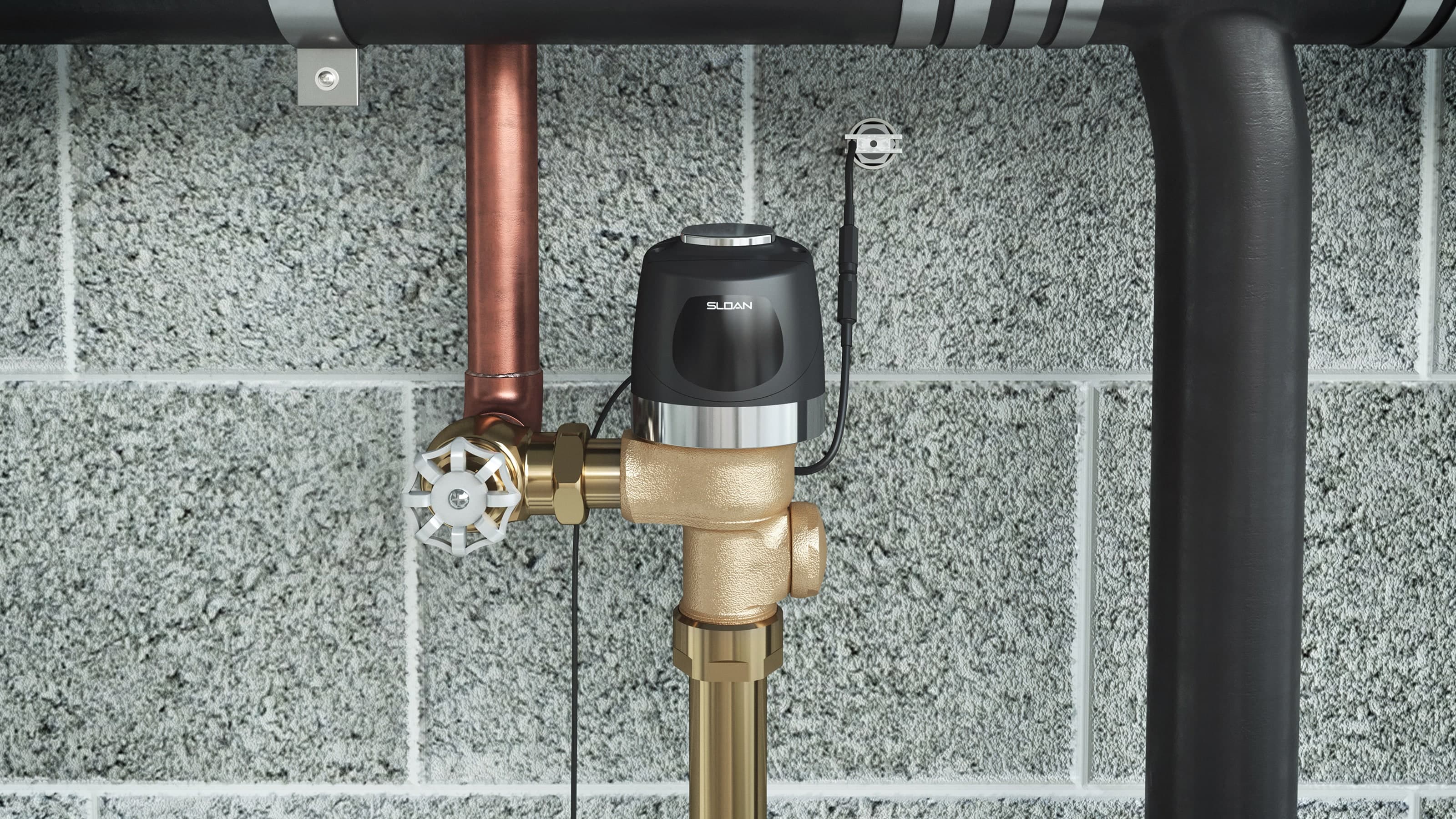 Brass valve assembly with a black actuator and manual wheel handle, connected to copper and black pipes against a concrete block wall.